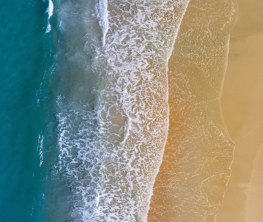 Imagen de una playa tomada con un plano aéreo, en el que se aprecian 3 colores: marrón de la arena, azul turquesa del agua y, en el centro, color blanco, del agua que llega a la costa.
